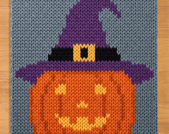 May include: A rectangular knitted blanket featuring a gray background with a central orange pumpkin wearing a purple witch hat. The hat includes a black band and a yellow buckle. The pumpkin has a yellow jack-o'-lantern face with eyes and a mouth.