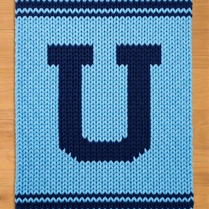 May include: A knitted blanket in shades of blue, featuring a large letter "U" in navy blue against a lighter blue background. The blanket has navy blue stripes at the top and bottom, creating a simple, yet bold design. The texture is chunky and cozy.