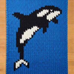 Puede incluir: Una manta azul tejida con un diseño de orca en blanco y negro. La orca se representa con sus distintivas marcas blancas y negras, sobre un fondo azul liso. La manta tiene una apariencia texturizada y tejida.