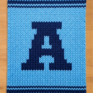 May include: A knitted blanket featuring a large, dark blue letter "A" on a light blue background. The blanket has a textured appearance with a striped border in dark blue. The design is simple and bold, suitable for home decor.