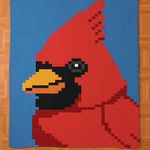 May include: A knitted blanket featuring a pixelated image of a red cardinal bird against a blue background. The bird has a black face, a yellow beak, and a white eye. The blanket is made of soft, textured yarn.