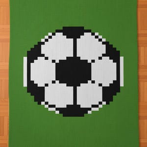 Puede incluir: Una manta verde tejida con un diseño de balón de fútbol pixelado en blanco y negro. La manta tiene forma rectangular y está hecha de un material suave y texturizado. El balón de fútbol está centrado en la manta.