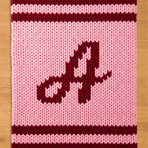 Puede incluir: Una manta de punto en tonos rosa y burdeos. La manta presenta una gran letra "A" en burdeos en el centro. Los bordes superior e inferior tienen rayas burdeos, y el resto de la manta es rosa.