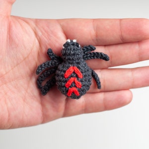May include: A small crocheted spider with a black body and red markings on its back. The spider is sitting on a person's hand.