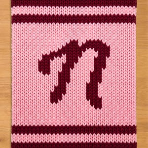 Könnte beinhalten: Ein gestricktes Quadrat in Rosa- und Burgundertönen. Das Mittelfeld ist hellrosa, mit einem burgunderfarbenen Buchstaben "M" in der Mitte. Die oberen und unteren Ränder sind burgunderfarben, und es gibt burgunderfarbene Streifen über und unter dem Buchstaben.
