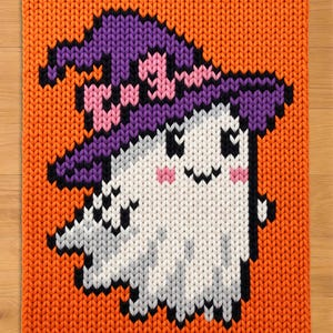 Può includere: Una coperta lavorata a maglia rettangolare con uno sfondo arancione vivace e un motivo centrale raffigurante un fantasma bianco che indossa un cappello da strega viola con dettagli rosa. Il fantasma ha occhi neri, bocca nera e guance rosa.