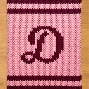 Puede incluir: Una alfombra rectangular tejida en tonos rosa y burdeos. La alfombra presenta una gran letra "D" en burdeos en el centro. Los bordes superior e inferior, junto con dos rayas, son burdeos. El fondo es rosa claro.