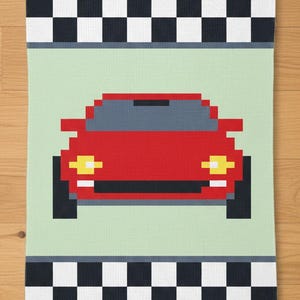 May include: A red pixelated car design on a light green background, with a black and white checkered pattern at the top and bottom. The car has yellow headlights and a gray roof. This design is likely for a kitchen towel or similar item.