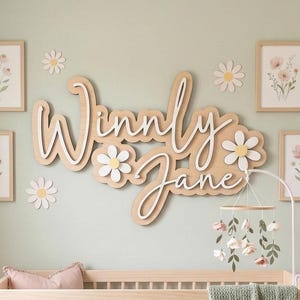 May include: A wooden wall sign featuring the name Winny Jane in a layered script font, accented with white and yellow daisy cutouts. The display is surrounded by four framed floral prints and small daisy wall decals above a wooden crib.