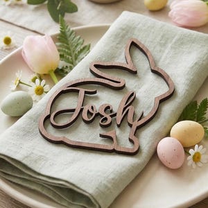 May include: A wooden bunny place card with the name "Josh" on a plate with a green napkin. The plate is decorated with pastel Easter eggs, flowers, and greenery, creating a festive table setting for Easter.