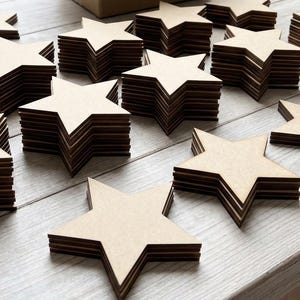 May include: Stacks of unfinished wooden star cutouts. Each star is composed of multiple layers, creating a dimensional effect. The stars are arranged on a light-colored wooden surface, with a cardboard box in the background.