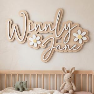 May include: A wall-mounted wooden sign featuring the name Winnly Jane in white cursive lettering layered over a light wood-toned backing. The design includes two white daisy flowers with yellow centers integrated into the layered wooden structure.