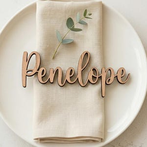 May include: A wooden name tag that reads "Penelope" in cursive script, placed on a folded beige linen napkin. The napkin rests on a white plate, with a sprig of greenery and a gold fork visible.
