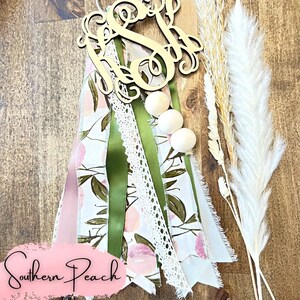 May include: A wooden monogram with the letter "S" in cursive, surrounded by a variety of ribbons and lace. The ribbons are in shades of pink, green, and white, and the lace is white. There are three wooden beads in the center of the image. The background is a wooden surface. The text "Southern Peach" is visible in the lower left corner of the image.