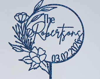 May include: A blue glitter cake topper with the words "The Robertsons" and the date "03.02.2026" inside a circular frame. Floral and leaf accents surround the frame, adding a decorative touch to this wedding cake decoration.