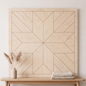 May include: A large, light-colored wooden wall art piece with a geometric star design. The artwork is displayed on a wooden table with a vase of dried plants and folded towels. The star pattern is made of dark brown lines.