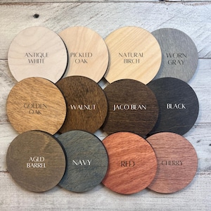May include: Twelve circular wooden discs arranged in a grid, each featuring a distinct wood stain color and a printed label. The labels read Antique White, Pickled Oak, Natural Birch, Worn Gray, Golden Oak, Walnut, Jaco Bean, Black, Aged Barrel, Navy, Red, and Cherry.