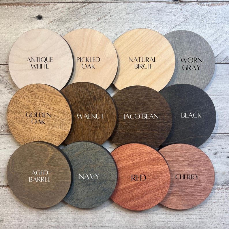 May include: Twelve circular wooden discs arranged in a grid, each featuring a distinct wood stain color and a printed label. The labels read Antique White, Pickled Oak, Natural Birch, Worn Gray, Golden Oak, Walnut, Jaco Bean, Black, Aged Barrel, Navy, Red, and Cherry.