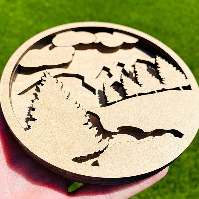 May include: A wooden wall decor with a cutout design of a mountain range and trees. The design is in a circular shape and has a light brown color.