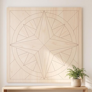 May include: A large, square wooden wall art piece featuring a compass rose design. The artwork is made of light-colored wood with black lines outlining the compass rose and geometric shapes. A small potted plant sits below the artwork.