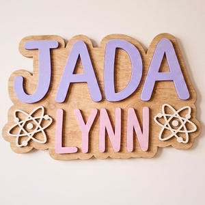 May include: Wooden sign with the name "JADA LYNN" in lavender and pink letters. The letters are mounted on a light brown wooden base with a cloud-like shape. Two white atom symbols flank the name.
