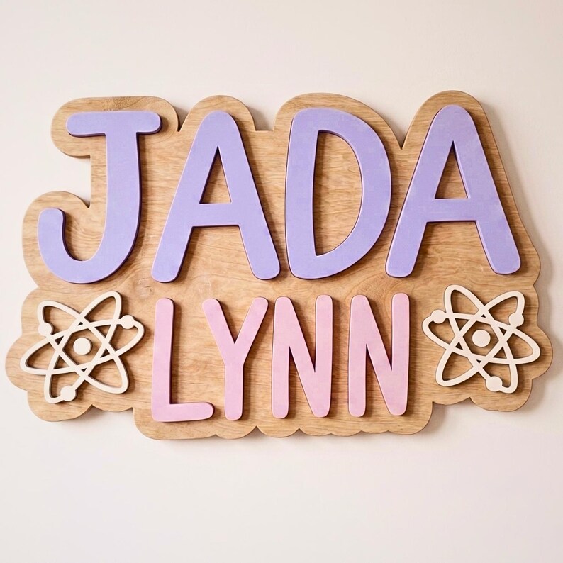 May include: Wooden sign with the name "JADA LYNN" in lavender and pink letters. The letters are mounted on a light brown wooden base with a cloud-like shape. Two white atom symbols flank the name.