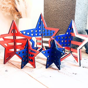May include: Five wooden stars painted in red, white, and blue with a star pattern. The stars are layered and create a 3D effect.