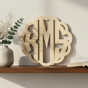 May include: A wooden monogram with the letters R, M, and S in a decorative, scalloped frame. The natural wood-toned monogram is displayed on a shelf with a vase of greenery and an open book.