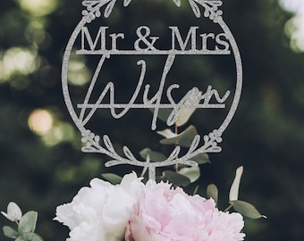 May include: Silver glitter cake topper with a floral wreath design. The topper says "Mr. & Mrs. Wilson" in a script font.