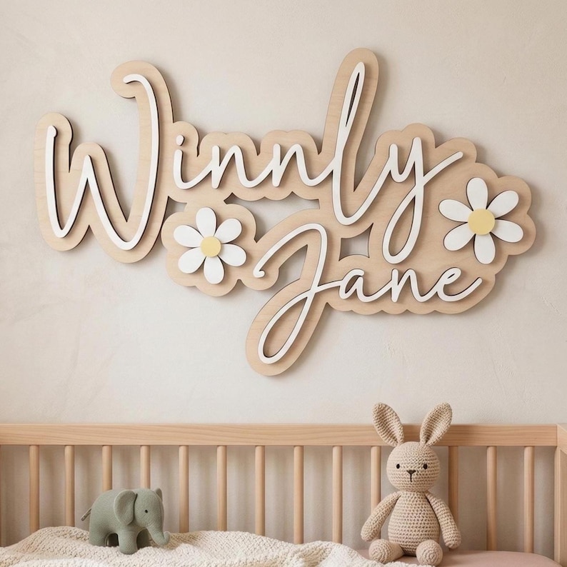 May include: A wall-mounted wooden sign featuring the name Winnly Jane in white cursive lettering layered over a light wood-toned backing. The design includes two white daisy flowers with yellow centers integrated into the layered wooden structure.