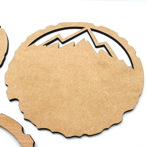 May include: A brown wooden craft blank with a black outline of a mountain range. The blank is round and has a rough texture.