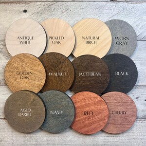 May include: A collection of twelve round wooden samples, each displaying a different stain color. Colors include Antique White, Pickled Oak, Natural Birch, Worn Gray, Golden Oak, Walnut, Jaco Bean, Black, Aged Barrel, Navy, Red, and Cherry.