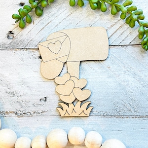 May include: Wooden cutout of a mailbox with a heart-shaped envelope sticking out of the top. The mailbox is sitting on a bed of grass with hearts around it.