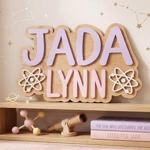 May include: A wooden name sign with the name "JADA LYNN" in lavender and pink, with atom symbols. The sign is displayed above a wooden shelf with books and a toy telescope. The background features constellation decals.