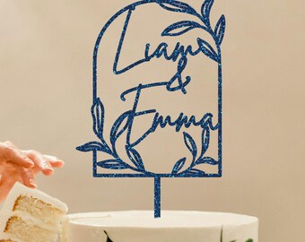 May include: Blue glitter wedding cake topper with a floral arch design and the names "Liam & Emma" written in cursive.