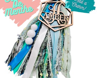 May include: A wooden charm with the name "AMBER" in a geometric shape, with a blue and green tassel. The charm has a hole at the top for adding a charm.