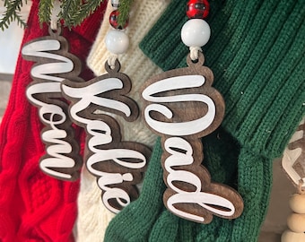 May include: Three personalized Christmas stockings with wooden ornaments that say "Mom", "Dad", and "Katie". The stockings are red, white, and green.