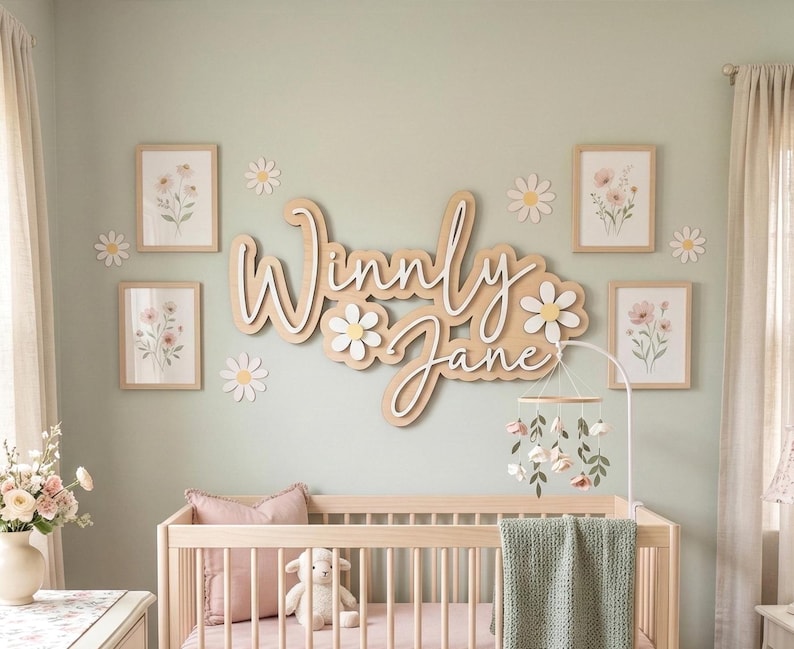 May include: A wooden wall sign featuring the name Winny Jane in a layered script font, accented with white and yellow daisy cutouts. The display is surrounded by four framed floral prints and small daisy wall decals above a wooden crib.