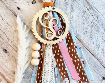 May include: A wooden monogrammed wall hanging with a scalloped edge, featuring the letter "L". It includes fabric strips in blue, brown, and pink, with lace and wooden beads. A metal ring is at the top.