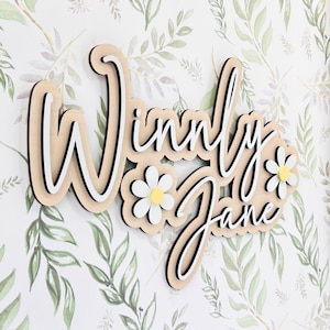 May include: A layered wooden wall sign featuring the name Winny Jane in a cursive script font. The design includes white and black layered lettering with two white daisy flowers with yellow centers integrated into the wooden name plaque.