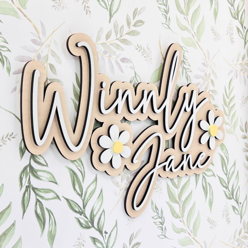 May include: A layered wooden wall sign featuring the name Winny Jane in a cursive script font. The design includes white and black layered lettering with two white daisy flowers with yellow centers integrated into the wooden name plaque.