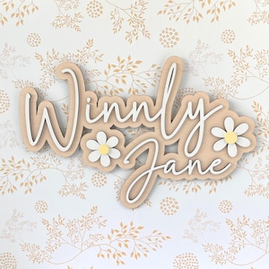 May include: A custom wooden wall sign featuring the name Winnly Jane in white script lettering layered over a light wood backing. The design includes two white daisy flowers with yellow centers positioned on the left and right sides of the text.