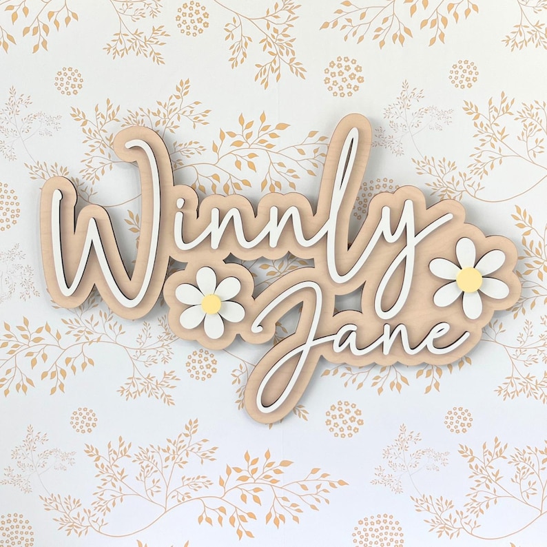 May include: A custom wooden wall sign featuring the name Winnly Jane in white script lettering layered over a light wood backing. The design includes two white daisy flowers with yellow centers positioned on the left and right sides of the text.