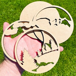May include: Three unfinished wooden circles with various nature-themed cutouts. The top circle has a mountain range, the middle circle has a cloud, and the bottom circle has a forest.