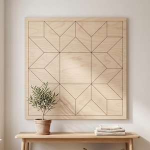 May include: A large, square wooden wall art piece with a geometric star pattern etched into the light-colored wood. The artwork is displayed above a wooden console table, with a small potted plant and folded linens.