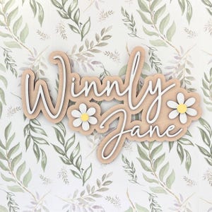 May include: A custom wooden wall sign featuring the name Winnly Jane in white script lettering layered over a light wood-toned backing. The design includes two white daisy flowers with yellow centers positioned on the left and right sides.
