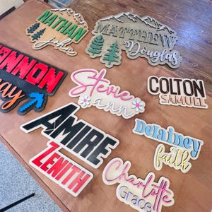 May include: A collection of custom laser-cut wooden name signs featuring various fonts, colors, and layered designs. The signs display names including Nathan John, Matthew Douglas, Cannon, Stevie Ann, Colton Samuel, Amire Zenith, Delainey Faith, and Charlotte Grace.