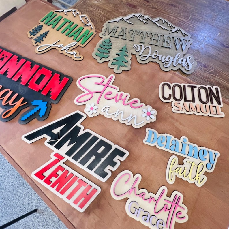 May include: A collection of custom laser-cut wooden name signs featuring various fonts, colors, and layered designs. The signs display names including Nathan John, Matthew Douglas, Cannon, Stevie Ann, Colton Samuel, Amire Zenith, Delainey Faith, and Charlotte Grace.
