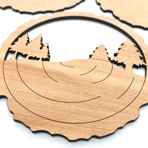 May include: A wooden coaster with a circular design featuring a forest scene with trees and a hill. The coaster has a natural wood grain and a dark brown border.
