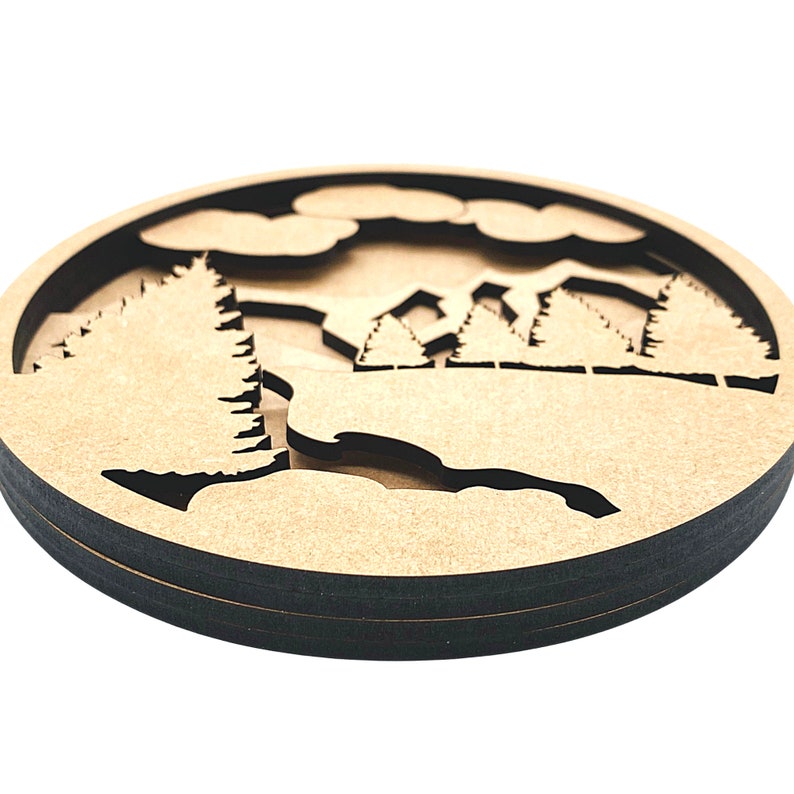 May include: A wooden wall hanging with a layered design of a forest scene. The design features trees, clouds, and a mountain range. The wall hanging is circular and has a dark brown border.
